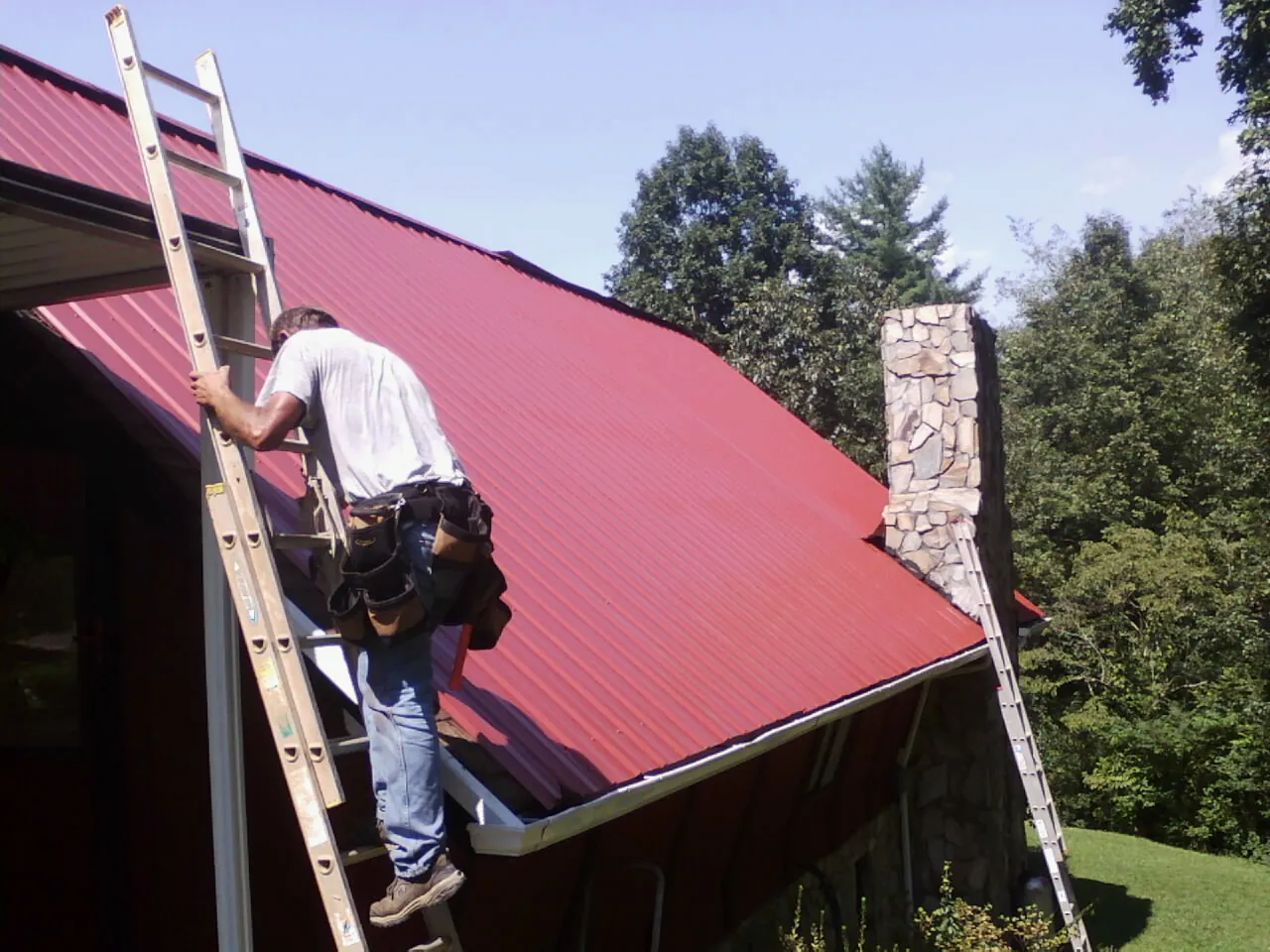 Roofer on a ladder beside a red metal roof and stone chimney — Soffit & Fascia Repair by Roofing Brothers in Jerseyville